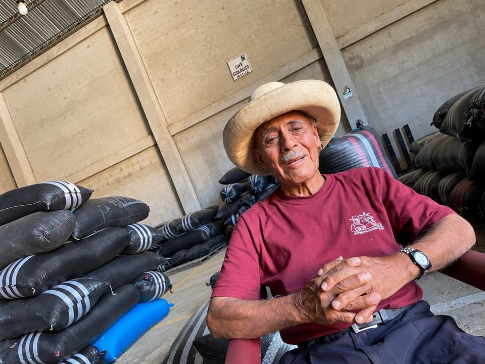 Socio fundador de APROECO con sombrero de paja, sonriente y sentado junto a sacos de café en el almacén.
