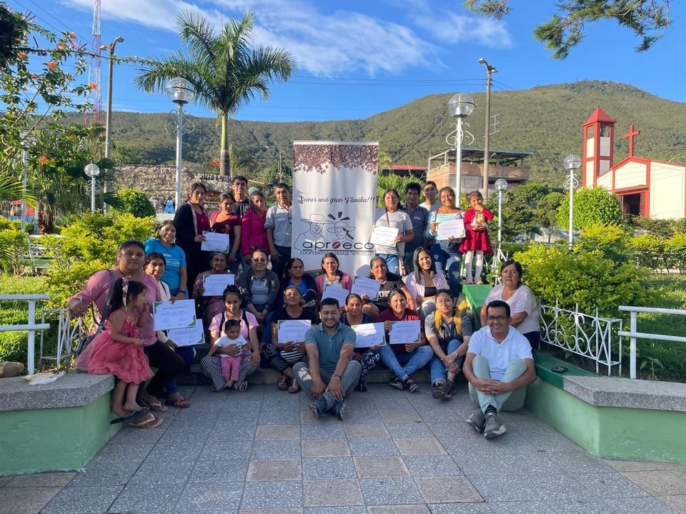 Socias y socios de APROECO con sus certificados, reunidos en la plaza junto al banner «Somos una gran familia».