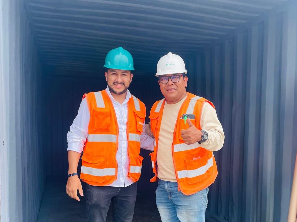 Dos integrantes del equipo con casco y chaleco frente a un contenedor de exportación.